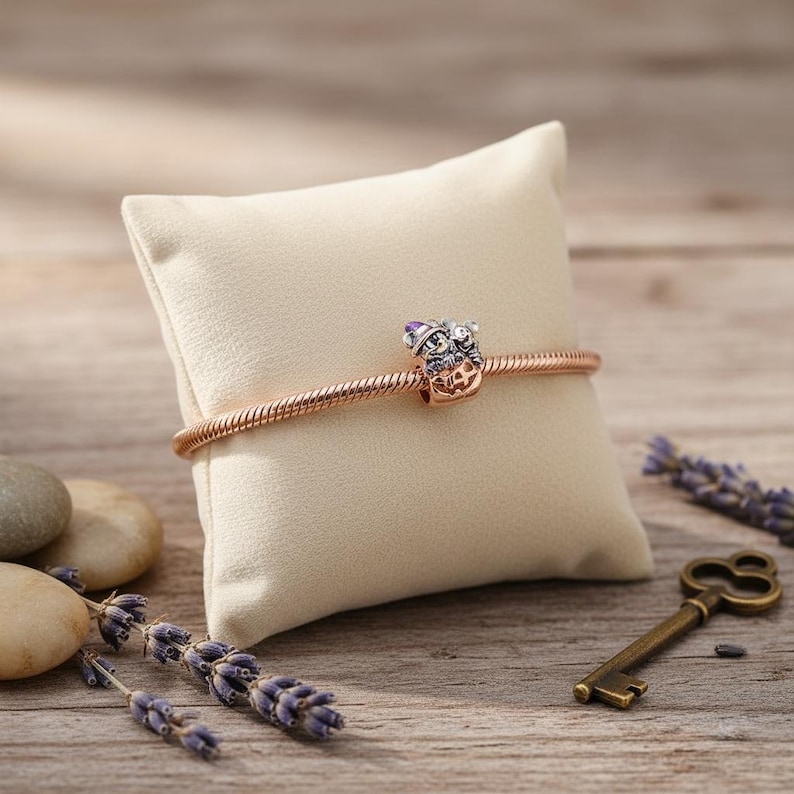 May include: A rose gold bracelet with a charm featuring a crown and purple accents, displayed on a cream-colored pillow. The bracelet is set against a wooden background with lavender sprigs and a vintage key.