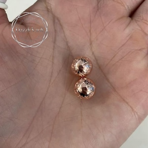 May include: Two rose gold spherical beads with intricate floral designs. The beads are displayed on a light background. The beads have a shiny, reflective surface. The beads are likely jewelry components.