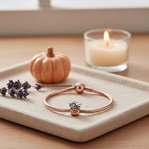 May include: A rose gold charm bracelet with a decorative charm, a small orange pumpkin, a lit candle, and lavender sprigs on a beige tray. The charm has a silver and gold design.