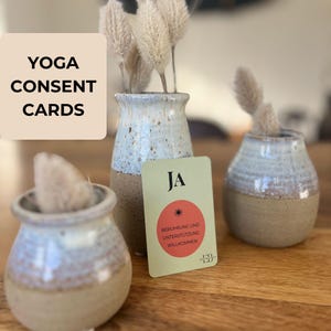 Könnte beinhalten: Das Bild zeigt Yoga-Einverständniskarten mit dem Text "YOGA CONSENT CARDS". Eine Karte mit dem Wort "JA" ist zu sehen, zusammen mit einem roten Kreis und deutschem Text. Die Karten werden in der Nähe kleiner Keramikvasen mit getrockneten Pflanzen präsentiert.