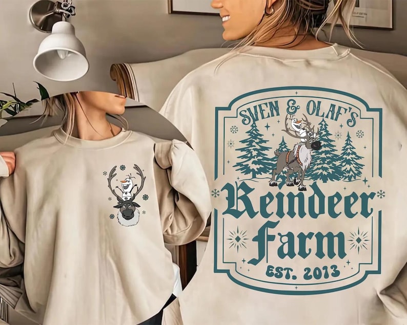 Pode incluir: Camisola bege com um desenho de Sven e Olaf de Frozen. A frente tem uma pequena imagem de Sven com Olaf em seus chifres. A parte de tr&aacute;s tem um design maior com o texto "Sven & Olaf's Reindeer Farm EST. 2013".