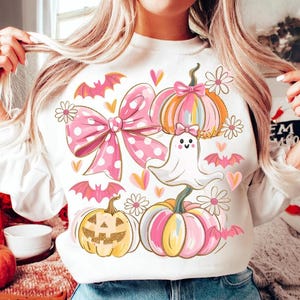 May include: White sweatshirt featuring a Halloween-themed design. The graphic includes a large pink polka-dot bow, pumpkins, bats, flowers, hearts, and a friendly ghost. The pumpkins are in shades of pink, orange, and yellow.