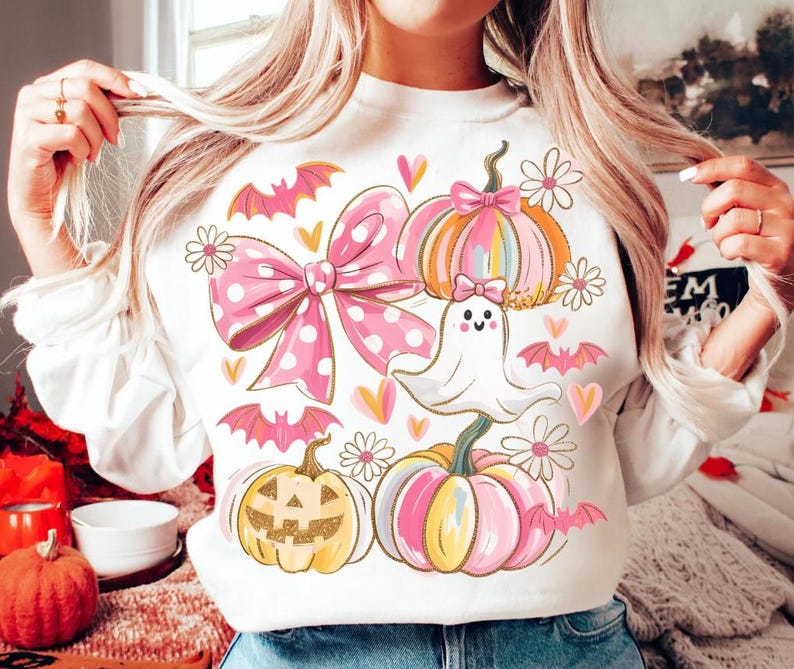 May include: White sweatshirt featuring a Halloween-themed design. The graphic includes a large pink polka-dot bow, pumpkins, bats, flowers, hearts, and a friendly ghost. The pumpkins are in shades of pink, orange, and yellow.