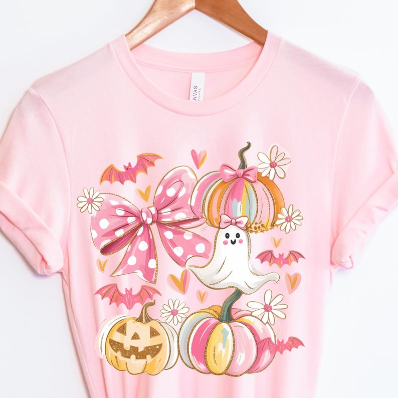 May include: Pink t-shirt featuring a Halloween-themed design. The graphic includes a large pink polka-dot bow, pumpkins, a ghost, bats, and daisies. The pumpkins are in shades of pink, orange, and yellow.