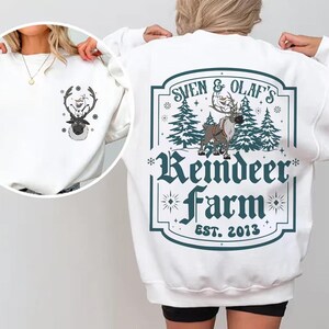 Pode incluir: Camisola branca com um desenho de rena e boneco de neve. A parte de tr&aacute;s apresenta um design turquesa e branco que diz "Sven & Olaf's Reindeer Farm EST. 2013". A frente tem um desenho menor da rena e do boneco de neve.