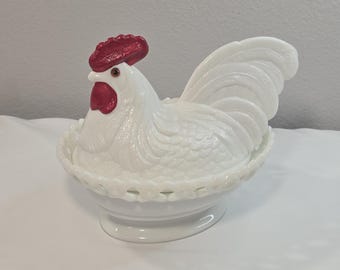 Puede incluir: Un plato de vidrio lechoso blanco con forma de gallina sobre un nido, con una tapa de gallo detallada que tiene una cresta y barbillas rojas. La base del plato presenta un patrón de celosía decorativo en el borde y plumas texturizadas.
