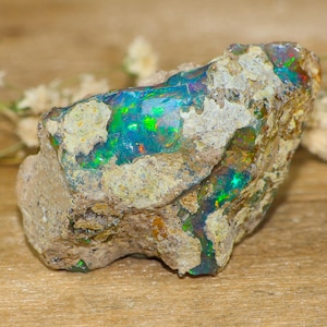 May include: A raw, unpolished opal specimen featuring a rough, sandy-coloured rock matrix embedded with vibrant, iridescent flashes of neon blue, green, and subtle orange light. The stone displays a natural, irregular shape with deep crevices.