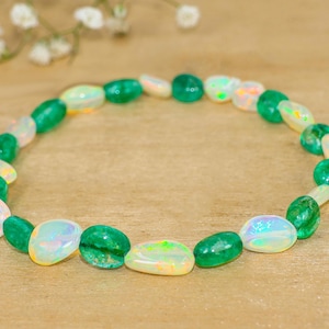Könnte beinhalten: Ein Armband mit abwechselnd grünen und schillernden Opalsteinen. Die oval geformten Perlen sind aufgereiht und bilden ein kreisförmiges Design. Das Armband wird auf einer hellbraunen Oberfläche mit kleinen weißen Blumen im Hintergrund präsentiert.