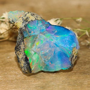 Puede incluir: Una piedra preciosa de ópalo en bruto que presenta un vibrante juego de colores con destellos dominantes en azul eléctrico, verde y naranja sutil. La piedra tiene un exterior de roca matriz rugoso, oscuro y texturizado en el lado izquierdo y una cara cristalina pulida.