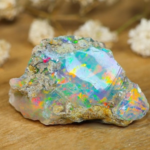 Puede incluir: Una piedra preciosa de ópalo en bruto y sin pulir que presenta una base translúcida de color blanco lechoso con destellos vibrantes de color iridiscente, incluyendo verde neón, naranja brillante y azul eléctrico, incrustada en una matriz rocosa rugosa.
