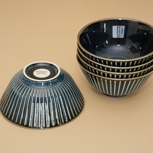 以下が含まれることがあります： 縦の白いストライプが入った、濃いネイビーブルーのセラミックボウル5個セット。1つのボウルは裏返しになっており、白い縁とテキストが表示されています。ボウルは光沢のある仕上がりで、金色の縁があります。