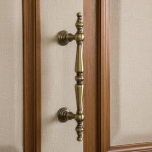 Op de afbeelding: Een bronzen deurkruk met een sierlijk ontwerp. De handgreep is verticaal georiënteerd en heeft decoratieve details over de gehele lengte, met een afgeronde boven- en onderkant. Gemonteerd op een lichtgekleurd, getextureerd oppervlak, omlijst door een houten deur.