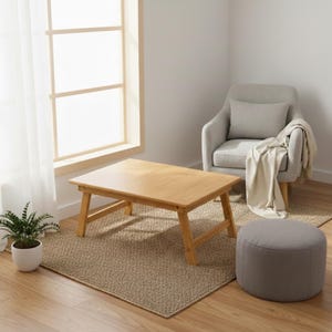 May include: A light brown, rectangular bamboo laptop table with folding legs. The table is set on a woven beige rug in a well-lit room. A gray armchair with a pillow and blanket is in the background, along with a small gray ottoman and a potted plant.