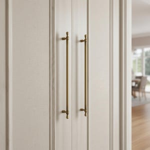 May include: Close-up of a pair of cabinet doors with long, brass-colored handles. The handles are vertically oriented and have a simple, elegant design. The doors are a light cream color with a subtle textured pattern.