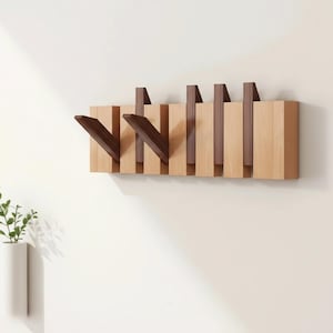 Op de afbeelding: Een houten wandkapstok met een modern design. Het heeft een reeks verticale houten latten in lichte en donkere bruintinten. Twee van de latten hebben schuine haken die naar buiten uitsteken. De kapstok is aan een witte muur bevestigd.