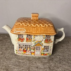 May include: A ceramic teapot in the shape of a cottage with a thatched roof. The cream-coloured teapot has a painted facade of a candy shop, with windows, flowers, and the text "Ye Olde Candy Shoppe."