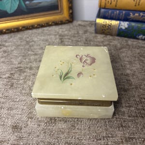 May include: A square, pale green stone box with a hinged lid featuring a painted floral design of a purple flower and green stems. The box has a metallic gold-toned band around the centre where the lid meets the base, providing a contrast to the smooth, translucent stone material of the container.