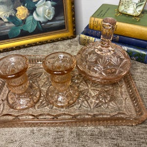 May include: A pink glass vanity set comprising a tray, two candle holders, and a lidded jar. The set features a starburst design and is presented on a textured surface. A framed painting and books are in the background.