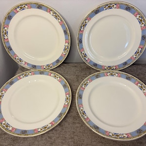 May include: A set of four round ceramic dinner plates with a cream-coloured centre and a decorative border. The rim features a light blue patterned band accented with small pink floral clusters, gold-toned filigree, and dark blue circular motifs.