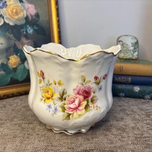 May include: A white porcelain planter with a scalloped rim and gold trim. The exterior features a floral pattern with yellow and pink roses, green leaves, and small blue flowers. The body has a textured, wavy surface and a wide, open top.