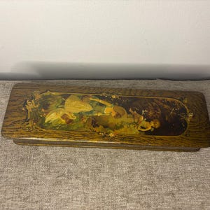 May include: A rectangular wooden box with a painted lid. The painting features figures in a landscape with yellow, green, and brown tones. The box has a dark wood grain finish, typical of antique items.