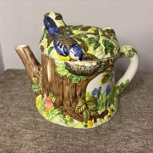 May include: A decorative ceramic teapot in the shape of a tree trunk, with a blue and yellow bird on the lid. The teapot is adorned with green foliage, flowers, and a nest. The handle and spout are crafted to mimic tree branches.