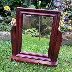 May include: A dark mahogany-stained wooden mirror with a rectangular frame. The mirror is supported by angled side supports and a base. The mirror reflects a garden scene. The frame and supports are a rich, reddish-brown colour.