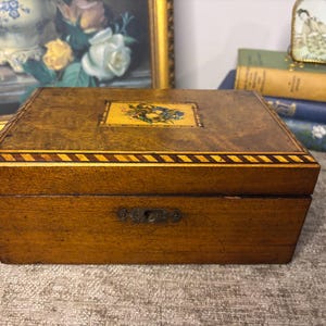 May include: A rectangular wooden box with a medium brown finish, featuring a central rectangular inlay depicting a floral arrangement. The lid is bordered by a dark and light wood striped pattern, and the front includes a metal keyhole escutcheon.