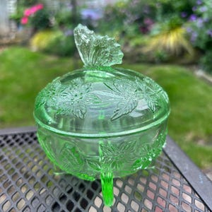 May include: A green glass candy dish with a lid, featuring a butterfly handle and floral patterns. The dish is round with three small feet. The background shows a garden with green grass and flowers.