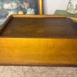 May include: A rectangular wooden box with a dark brown stained lid and a lighter honey-coloured front panel. The box features a flat top surface and a base with a slightly wider edge, showcasing a natural wood grain texture throughout the construction.