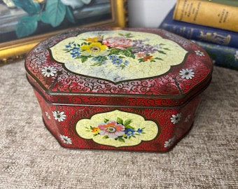 May include: An octagonal metal storage tin featuring a red background with intricate black scrollwork and white daisy accents. The lid and front panel display a cream-colored central cartouche decorated with a colorful floral bouquet of roses.