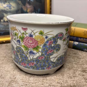 May include: A white ceramic planter featuring a colourful floral pattern with pink and yellow chrysanthemums and green leaves, accented by a blue swirling wave design around the base. The rim has a thin silver metallic trim around the top edge.