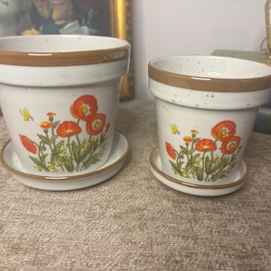 May include: Two ceramic flower pots with matching saucers, featuring a speckled off-white finish, a brown rim, and a vibrant orange poppy floral pattern. The pots are tapered with a wide opening and a sturdy base, designed for indoor plant display.