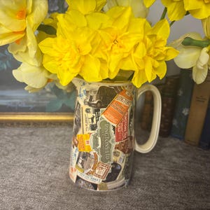 May include: A decorative jug, decorated with vintage labels, holding a bouquet of bright yellow daffodils. The jug has a white handle and is set against a backdrop of books and a framed picture.