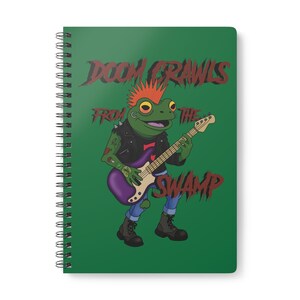 Peut inclure: Cahier à spirales avec une couverture verte. Il présente une grenouille de dessin animé jouant d'une guitare électrique violette, portant une veste en cuir noire, un jean bleu et des bottes. Le texte "DOOM CRAWLS FROM THE SWAMP" est écrit en rouge.