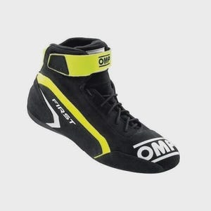 Op de afbeelding: Zwarte en gele raceschoen met het woord "FIRST" in het wit aan de zijkant. De schoen heeft een high-top ontwerp met een klittenband en het woord "OMP" in het wit. De schoen is gemaakt van su&egrave;de-achtig materiaal.