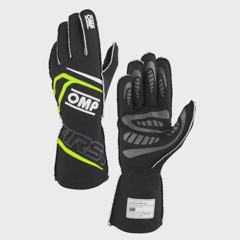 Op de afbeelding: Zwarte en neon groene racehandschoenen met het OMP-logo. De handschoenen hebben witte accenten en een label op de pols. Het woord "FIRST" staat in grote letters op de zijkant van de handschoenen.
