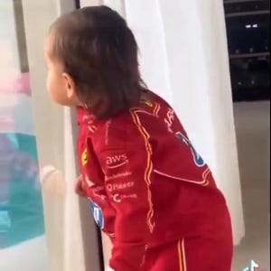 May include: A child wearing a red racing suit with white and yellow accents, including logos for "AWS", "Q-Pavar", and "C". The suit has a zipper closure and is paired with matching pants. The child is looking out a window.