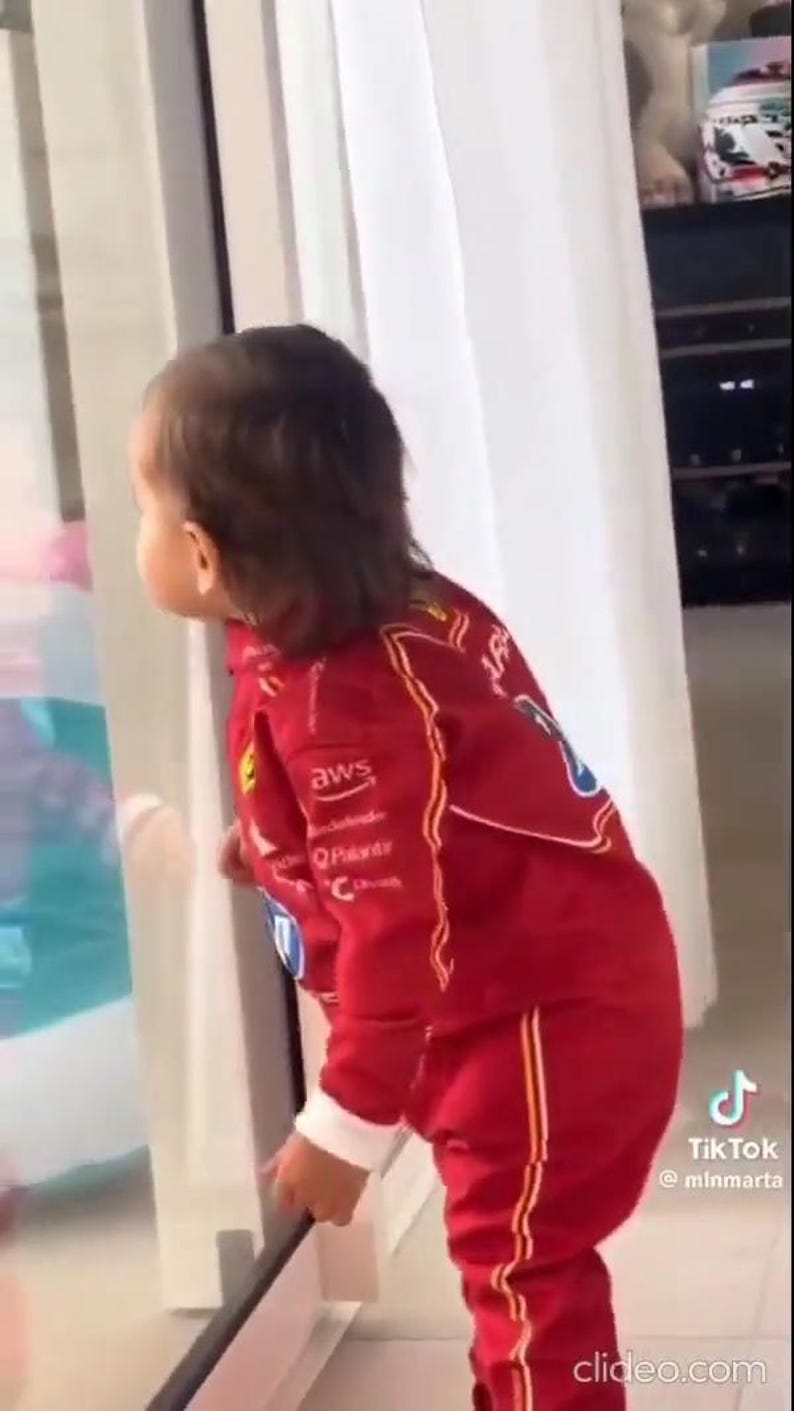 May include: A child wearing a red racing suit with white and yellow accents, including logos for "AWS", "Q-Pavar", and "C". The suit has a zipper closure and is paired with matching pants. The child is looking out a window.