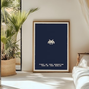 Puede incluir: Una impresión artística enmarcada con un fondo azul marino, que muestra un alienígena pixelado. El texto en la parte inferior dice "FIRST WE TAKE MANHATTAN THEN WE TAKE BERLIN". La obra de arte se exhibe en una habitación con luz natural.