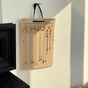 Könnte beinhalten: Ein beiger Schmuck-Organizer mit einem schwarzen Aufhängeband, montiert an einer weißen Wand. Der Organizer zeigt verschiedene goldene Halsketten und Ohrringe. Der Organizer hat kleine Haken und Löcher zum Aufhängen von Schmuck.