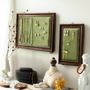 May include: Two framed jewelry organizers with green velvet backing and brown wooden frames hang on a white wall. The organizers display necklaces and earrings. A jewelry bust, small statues, and ring displays sit on a white surface.