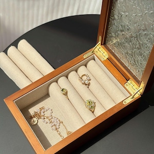 Op de afbeelding: Een houten sieradendoos met een glazen deksel, open om verschillende ringen en een ketting te laten zien. De ringen zijn onder meer een gouden ring met parels, een gouden ring met een groene steen en een gouden ring met gele stenen. De ketting is goud met parels.