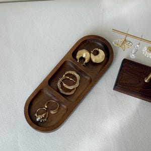 May include: A wooden jewelry tray with three compartments, holding gold hoop earrings, twisted hoop earrings, and a ring with pearls. The tray is a warm brown color and has a smooth finish.