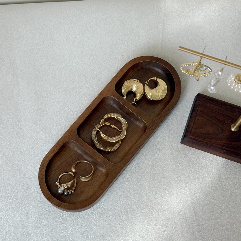 May include: A wooden jewelry tray with three compartments, holding gold hoop earrings, twisted hoop earrings, and a ring with pearls. The tray is a warm brown color and has a smooth finish.