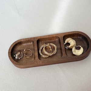 May include: A wooden jewelry tray with three compartments, holding various gold-toned earrings and rings. The earrings include pearl-studded hoops, a simple ear cuff, twisted hoops, and fan-shaped hoops. The tray is a warm brown color.