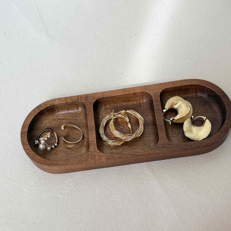 May include: A wooden jewelry tray with three compartments, holding various gold-toned earrings and rings. The earrings include pearl-studded hoops, a simple ear cuff, twisted hoops, and fan-shaped hoops. The tray is a warm brown color.