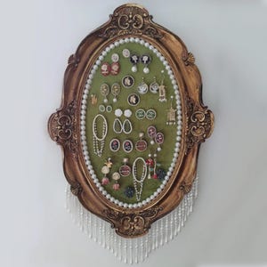 May include: An oval jewellery organiser with a gold-coloured frame. The interior has a green velvet backing and displays various earrings and necklaces. A pearl strand borders the green velvet, and a pearl fringe hangs from the bottom.