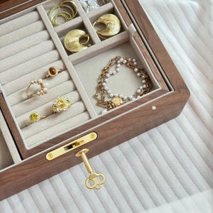 Puede incluir: Un joyero de madera oscura con interior de terciopelo beige que cuenta con ranuras para anillos y compartimentos abiertos. La caja contiene pendientes de aro dorados, anillos con piedras amarillas y blancas, y una pulsera de cadena dorada.