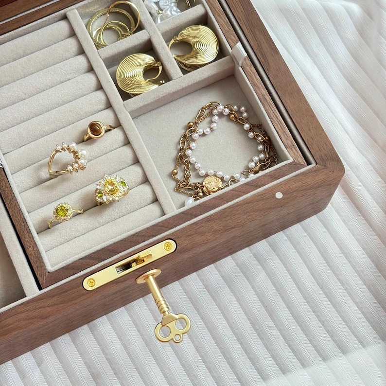 Puede incluir: Un joyero de madera oscura con interior de terciopelo beige que cuenta con ranuras para anillos y compartimentos abiertos. La caja contiene pendientes de aro dorados, anillos con piedras amarillas y blancas, y una pulsera de cadena dorada.